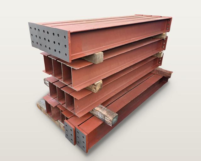 Pieces of spliced steel attic beams, painted red and stacked on timbers, with drilled end plates on their ends